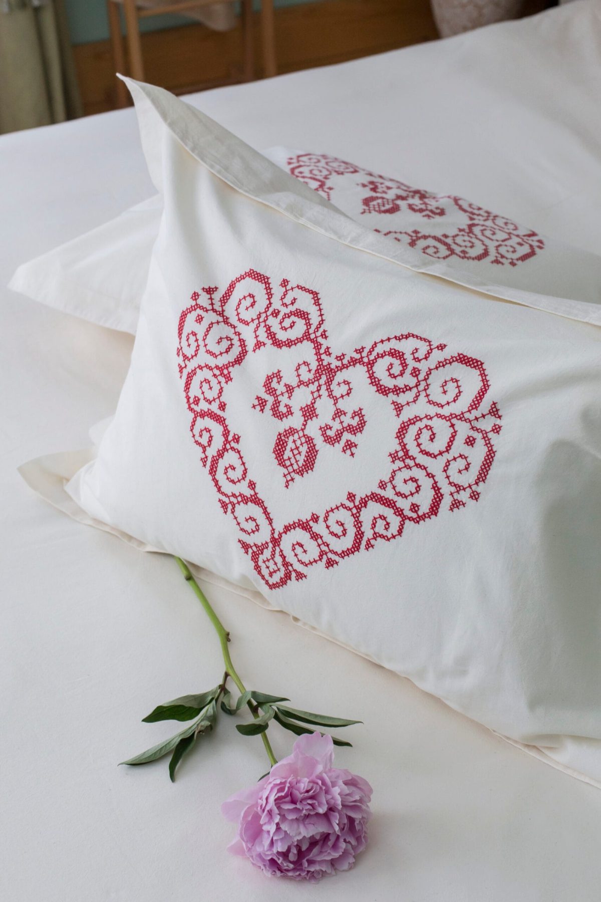 Two pillows in white cotton bed linen, with a heart embroidered in red thread and a purple flower